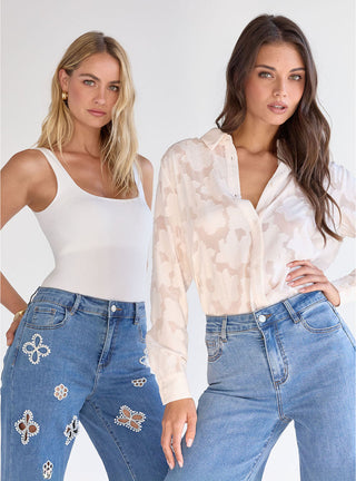 Two women wearing blue jeans and white tops stand side by side. One wears a sleeveless tank and the other a patterned button-up shirt. Both have loose hair and a neutral background behind them.