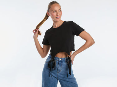A person with long braided hair, wearing a white sleeveless top with decorative pins and black leather pants, stands with hands in pockets against a plain background.