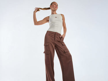 A person in a sleeveless black top and black high-waisted trousers with a double-buckled belt, standing against a light background. The person has their hands in their pockets.