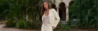 A woman with long dark hair wears a brown leather jacket with gold buttons, a beige top, and dark jeans with a gold chain belt, standing in front of a rustic wooden and metal background.