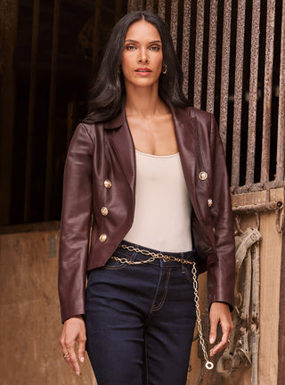 A woman with long dark hair wears a brown leather jacket with gold buttons, a beige top, and dark jeans with a gold chain belt, standing in front of a rustic wooden and metal background.