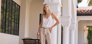 A woman with long blonde hair, wearing a white sleeveless top and patterned trousers, stands confidently by white pillars in an outdoor setting with Mediterranean-style architecture.
