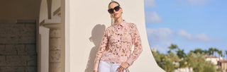 A woman wearing dark sunglasses, a pink lace blouse, and white pants stands against a light-colored, ornate building wall on a sunny day with a blue sky and palm trees in the background.