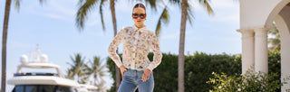 A woman poses confidently wearing a light pink floral blouse and pale pink pants with flower cutouts, holding a small pink handbag against a white background.