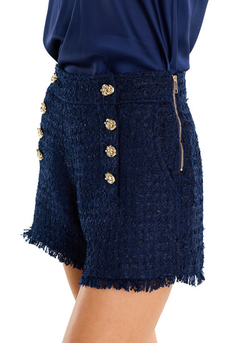 A person wearing Tamara Tweed Button Short - Navy, featuring textured navy tweed, gold decorative buttons, side zipper, and frayed hems with a matching navy top. Only the lower torso and upper thigh are visible.