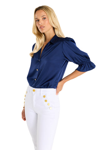 A woman in the Cindy Ruffle Sleeve Blouse - Navy, featuring shiny fabric and puffed three-quarter sleeves, pairs it with white high-waisted pants with gold buttons, standing against a plain white background.