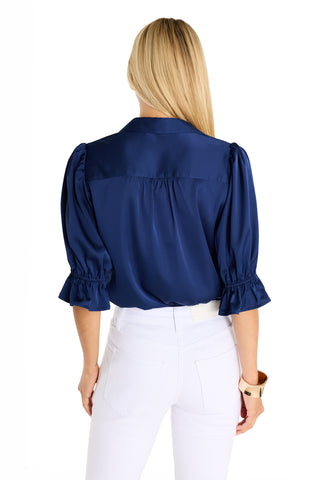 A woman with long blonde hair is seen from the back, wearing the Cindy Ruffle Sleeve Blouse in navy and white pants. She has a tan bracelet on her left wrist, set against a white background.