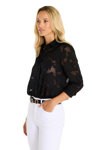 A woman with long blonde hair models the Stelle Button Up - Black, styled over a black bra with white pants and a black belt, standing against a white background.
