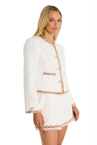 A woman wears the Joane Tweed Jacket - Pearl Multi, a white textured jacket and skirt suit with pearl buttons and light pink trim, standing at an angle against a plain white background.