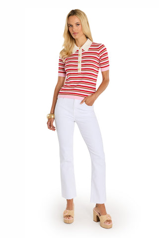 A woman stands confidently in the Leya Polo Shirt - Summer Multi, paired with white pants, beige platform sandals, and chunky bangles, with one hand in her pocket against a plain white background.