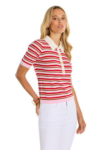 A woman with long blonde hair wears the Leya Polo Shirt - Summer Multi, featuring red and pink stripes, a white collar, and gold embossed buttons, paired with white pants against a white background.