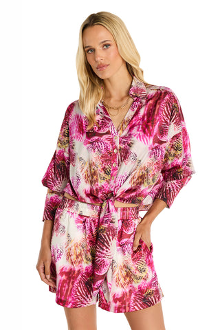 A woman with loose blonde hair, wearing the Cecilia Silk Tie-Front Blouse in Bright Orchid floral print, poses with hands in pockets against a white background.