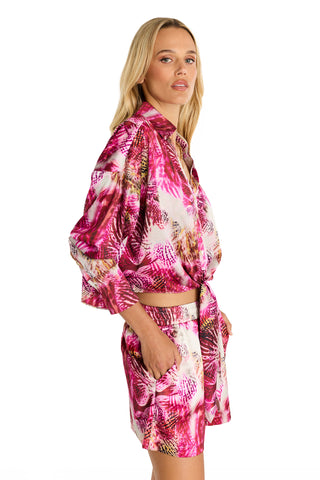 A woman poses with her hand in her pocket, wearing the Cecilia Silk Tie-Front Blouse in Bright Orchid—a vibrant pink, purple, and white floral orchid print—paired with matching shorts against a white background.