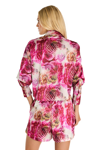 A woman stands with her back to the camera, wearing the Cecilia Silk Tie-Front Blouse in Bright Orchid, featuring a vibrant pink, white, and yellow abstract pattern. Her hands are in her pockets, and her blonde hair is loose.