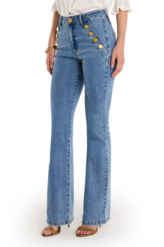 A person wears the Allegra Bootcut Jean - Medium Blue with gold hip detailing, a white blouse, tan heeled sandals, and a gold bracelet, standing against a white background.