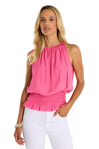 A woman with long blonde hair stands against a white background wearing the Betsy Halter Top in Petal Pink, featuring silky soft fabric, a gathered neckline, smocked waist, and paired with white pants.