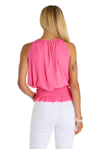 A woman with long blonde hair faces away, wearing the Betsy Halter Top in Petal Pink—silky, sleeveless, with adjustable ruching at the waist—and white pants. She has a tattoo on her upper left arm and a beaded bracelet on her right wrist.