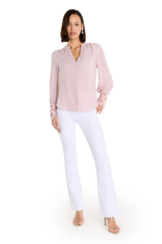 A woman with shoulder-length dark hair poses against a white background wearing the Blake Lace Sleeve Blouse in blush, styled with white flared pants and clear high-heeled sandals, her hand casually placed in her pocket.