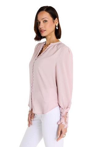 A woman with short dark hair models the Blake Lace Sleeve Blouse in blush, featuring lace cuffs, paired with white pants and pearl earrings. She poses three-quarters to the camera against a white background.
