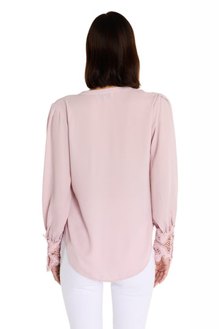 A woman with medium-length brown hair is seen from the back, wearing the Blake Lace Sleeve Blouse in blush with elegant lace cuffs and white pants against a white background.