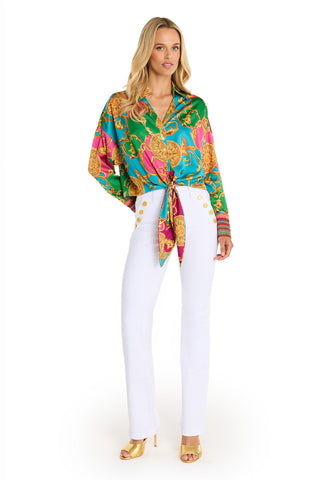 A woman poses against a white background wearing the Cecilia Silk Tie-Front Blouse - Dolce Sun, featuring green, pink, and gold patterns, paired with high-waisted white pants with gold buttons and gold high-heeled sandals.