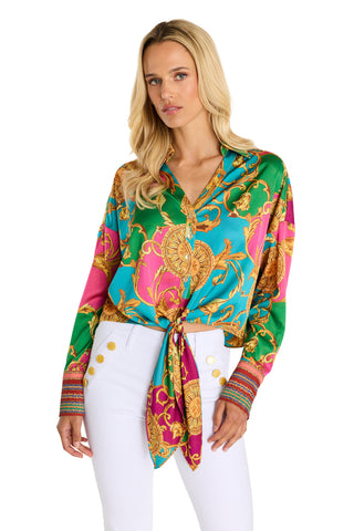 A woman with long blonde hair wears the Cecilia Silk Tie-Front Blouse - Dolce Sun, a vibrant limited edition top with gold baroque patterns in teal, pink, and green, paired with high-waisted white pants featuring gold buttons.