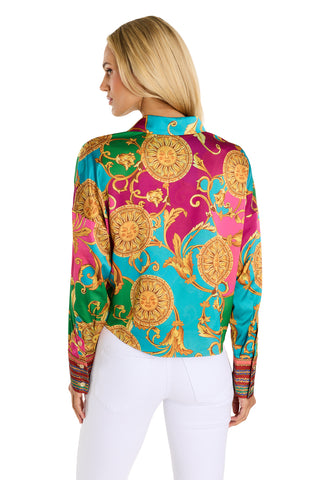 A woman with long blonde hair faces away, wearing the Cecilia Silk Tie-Front Blouse - Dolce Sun featuring gold sun and floral patterns, paired with fitted white pants. The background is plain white.