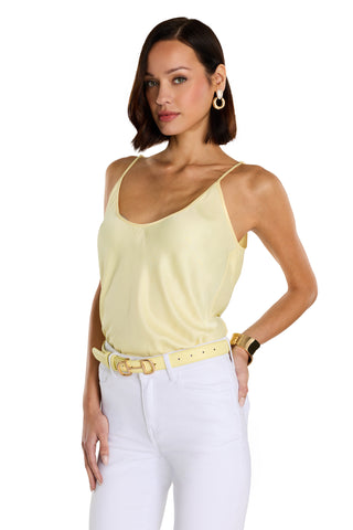 A woman with short brown hair wears a sunshine yellow Yara Silk Camisole, white pants, a yellow belt, gold hoop earrings, and a gold bracelet, standing with one hand on her hip against a white background.