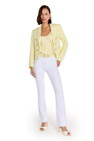 A woman with shoulder-length brown hair poses confidently against a white background, wearing the Gabrielle Double Breasted Blazer in Sunshine Yellow with a matching top, white pants, yellow belt, white heels, and gold earrings.