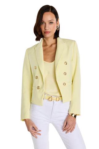 A woman with short brown hair wears the Gabrielle Double Breasted Blazer in Sunshine Yellow, featuring gold buttons and a tailored waist, over a matching top. She pairs it with white pants and gold accessories against a white background.