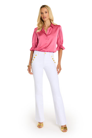 A woman with long blonde hair stands facing forward, wearing the Cindy Ruffle Sleeve Blouse in Petal Pink, white high-waisted pants with gold buttons, gold open-toe heels, and a white background accentuating her chic style.