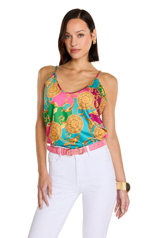 A woman with straight dark hair wears the Yara Silk Camisole - Dolce Sun, featuring gold designs and adjustable straps, paired with white pants and a pink belt. She poses with one hand in her pocket against a white background.