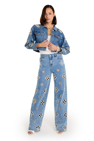 A woman wears the Kaia Pearl Jean Jacket in medium blue with matching wide-leg jeans featuring large floral cutouts, styled over a white top and heeled sandals, standing against a white background.