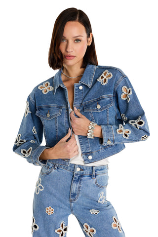 A woman with straight brown hair and a neutral expression poses with hands adjusting the Kaia Pearl Jean Jacket in medium blue, featuring white and brown flower-shaped embellishments and paired with matching jeans.