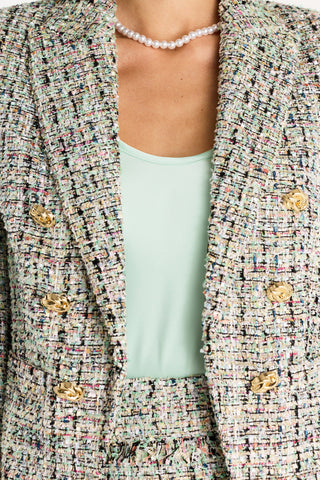 A person wears the Gabrielle Double Breasted Tweed Blazer - Teal Multi with gold buttons, a light green top, matching skirt, and a pearl necklace. The cropped image highlights the tailored waist, torso, and neck.