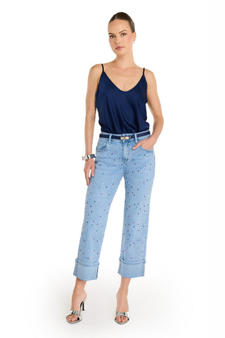 A woman stands confidently against a white background wearing the Ren Cropped Cuff Jean - Confetti, featuring four-way stretch denim with small colorful patterns, a navy blue sleeveless top, silver sandals, and a chunky bracelet.