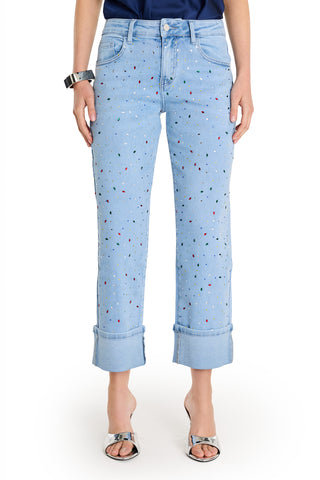 Wearing the Ren Cropped Cuff Jean - Confetti, featuring four-way stretch denim and colorful paint splatters, plus a dark blue top, silver bracelet, and open-toe silver heels, a person stands against a plain white background.