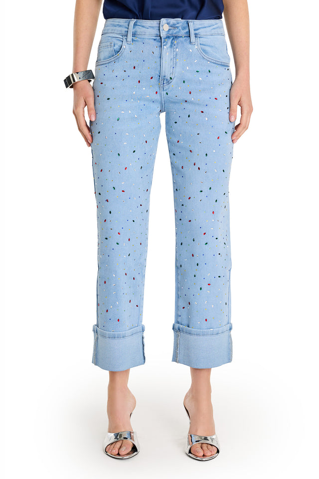 Wearing the Ren Cropped Cuff Jean - Confetti, featuring four-way stretch denim and colorful paint splatters, plus a dark blue top, silver bracelet, and open-toe silver heels, a person stands against a plain white background.