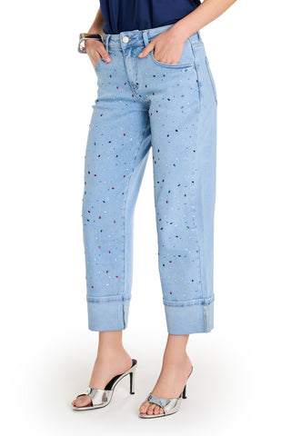 Wearing the Ren Cropped Cuff Jean - Confetti in four-way stretch denim with rainbow gem embellishments, a navy top, silver heeled sandals, and a chunky bracelet, a person stands hands in pockets.