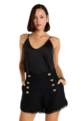 A woman with straight brown hair wears a black sleeveless satin top and the Tamara Tweed Button Short - Black with gold buttons, standing against a white background.