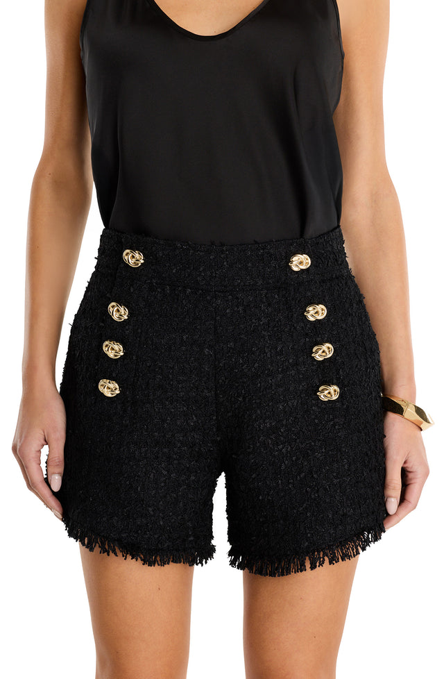 A woman wears a sleeveless black top and Tamara Tweed Button Short in black, featuring gold decorative buttons, frayed hems, and a gold side zipper. She accessorizes with a gold bracelet on her right wrist.