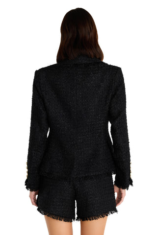 A woman with wavy brown hair is seen from the back wearing the Giselle Single Breasted Tweed Blazer in black, paired with matching black frayed-hem shorts. Gold buttons on the blazer sleeves add a chic accent.