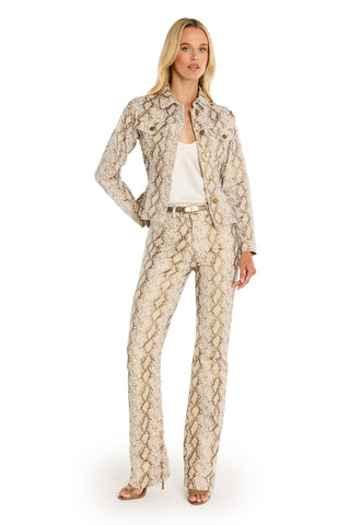 A woman poses with one hand on her hip against a white background, wearing the Bethanie Denim Jacket in Gold Python print over a white top, paired with matching pants and open-toe heels.
