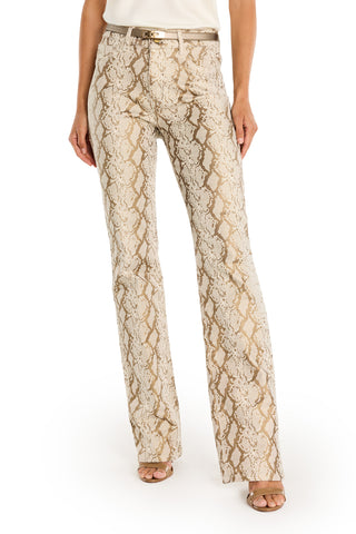 A person wearing the Aria Flare Jean - Gold Python, featuring a beige and gold snakeskin pattern, paired with a white sleeveless top, beige belt, and open-toe heels, stands against a white background.