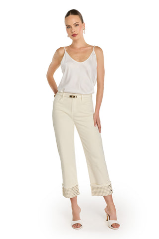 A woman poses confidently with her hand in her pocket, wearing the Ren Cropped Cuff Jean - Gold Mirror against a white background. She styles them with a white sleeveless top, white heels, and gold hoop earrings.