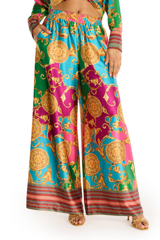 Wearing the Marcia Silk Pant - Dolce Sun, featuring an ornate gold pattern on vibrant blue, pink, and green silk, paired with gold sandals and a matching top partially visible—ideal for a chic summer print vibe.