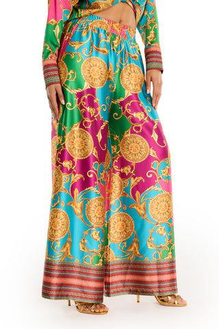 A woman wears the Marcia Silk Pant - Dolce Sun, designer high-waisted wide-leg pants with a vibrant baroque summer print in gold, pink, green, and turquoise, paired with gold heels.
