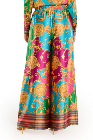 A person wearing the Marcia Silk Pant - Dolce Sun, featuring a colorful, ornate print with gold, turquoise, magenta, and green medallions and scrolls. These high-waisted wide-leg silk pants offer a loose fit for a bold summer look.