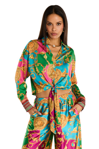 A woman confidently poses with hands in pockets, wearing the Cecilia Silk Tie-Front Blouse in Dolce Sun, featuring a vibrant baroque-style gold pattern on green, blue, and pink silk-like fabric paired with wide-leg pants.
