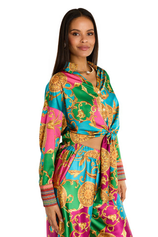 A woman stands against a white background wearing the Cecilia Silk Tie-Front Blouse - Dolce Sun, featuring bold gold, pink, green, and blue mosaic patterns. The blouse is styled at the waist, and she smiles gently.